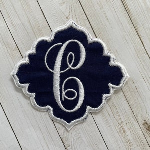 May include: Navy blue embroidered patch with a white scalloped edge and a white script letter "C" in the center. The patch has a decorative shape, suitable for personalization on clothing or accessories.