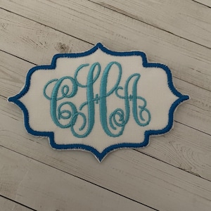 May include: Embroidered patch featuring a monogram in light blue cursive script. The monogram is set against a white background with a blue border. The patch has a decorative, scalloped edge.