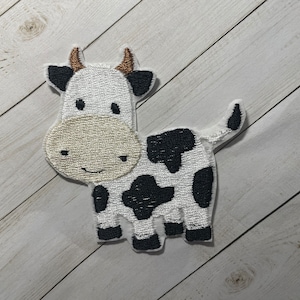 May include: A white and black embroidered cow patch with brown horns. The cow has a smiling face and black spots.
