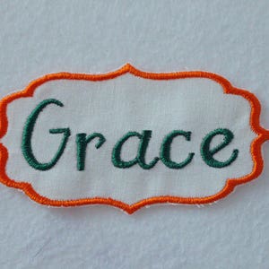 May include: Embroidered patch with the name "Grace" in green script. The name is centered on a white fabric, framed by an orange decorative border. The patch is likely for clothing or crafts.