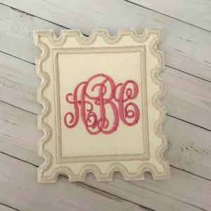 May include: A rectangular fabric patch shaped like a postage stamp with scalloped edges. The patch is off-white with a beige border and features a pink embroidered monogram of the letters ABC in a cursive script centered on the fabric surface.