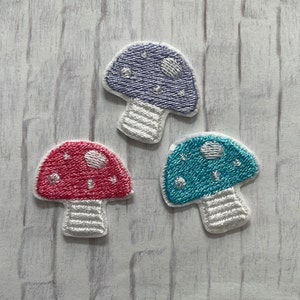 May include: Three embroidered mushroom patches in red, blue, and purple. Each mushroom has a white stem and a dotted cap. The patches are designed with a textured, stitched appearance, perfect for adding to clothing or accessories.