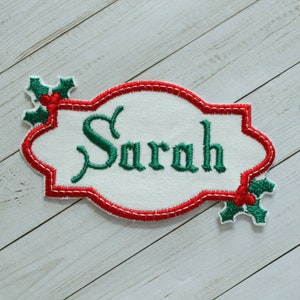 May include: Embroidered name patch with the name "Sarah" in green script. The patch has a white background with a red border and holly accents. The patch is approximately 4 inches wide and 2 inches tall.