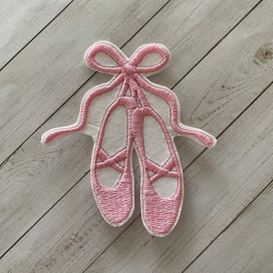 May include: Embroidered patch of pink ballet pointe shoes with a bow and ribbons. The patch is outlined in white and has a textured appearance. The design is on a light wood grain background.