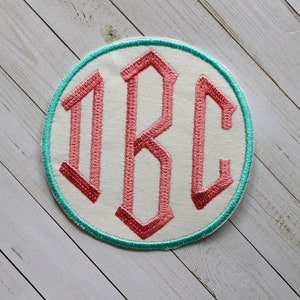 May include: Embroidered patch with the letters "JBC" in a pink, gothic-style font. The letters are on a white background, surrounded by a teal border and a white outer edge. The patch is circular.
