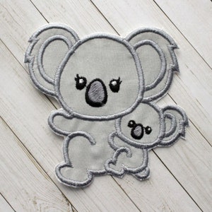 May include: A gray embroidered patch of a koala holding a smaller koala. The koalas have black eyes and noses.