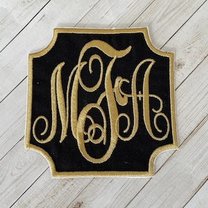 May include: A black fabric patch with a gold embroidered monogram of the letters "MGH" in a decorative script font. The patch has a scalloped edge and is surrounded by a gold border.
