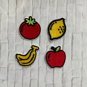 May include: Four embroidered fruit patches: a red tomato with green leaves, a yellow lemon with black details, a yellow banana, and a red apple with a green stem. Each patch has a white border and black outline.
