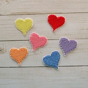 May include: Six embroidered heart patches in various colors: yellow, red, pink, orange, purple, and blue. Each heart has a white border and is stitched with a textured thread. The patches are arranged on a light wood-grain surface.