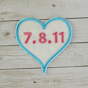 May include: A white heart-shaped patch with a blue outline. The patch has the text "7.8.11" embroidered in pink.