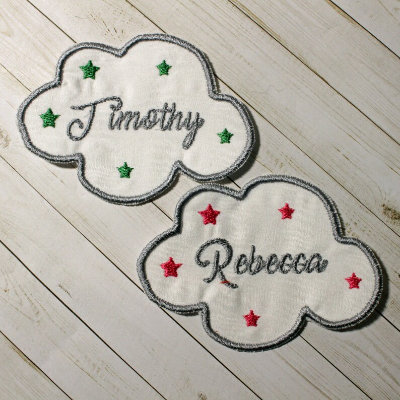 Pu&ograve; includere: Due toppe di tessuto a forma di nuvola bianca con nomi ricamati, "Timothy" e "Rebecca", e stelle verdi e rosse intorno ai nomi.