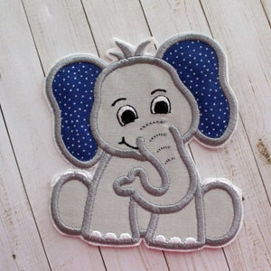 May include: A gray felt elephant applique with blue polka dot ears. The elephant is sitting with its trunk in its mouth and has a smiling face.