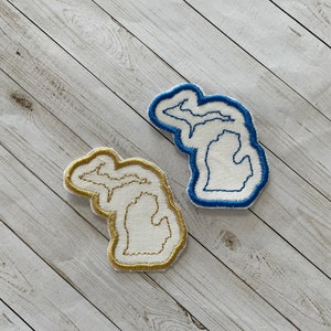 May include: Two embroidered patches in the shape of the state of Michigan. One patch is blue with white stitching, and the other is gold with white stitching.