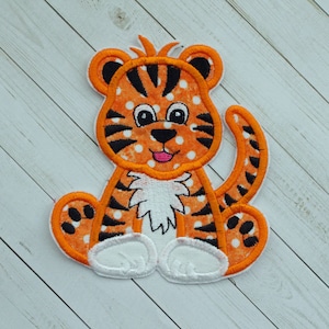 May include: A cute orange and black tiger applique with white polka dots and white paws. The tiger is sitting with its mouth open and tongue sticking out.
