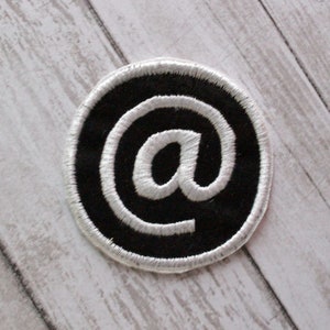 May include: Embroidered patch featuring a white "at" symbol on a black circular background. The patch has a white stitched border. The patch is placed on a light gray and white wooden surface.