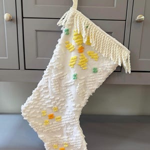 May include: White Christmas stocking with a textured pattern and fringe trim. The stocking is decorated with yellow, orange, and green embroidered floral designs. A cream-colored ribbon is attached for hanging.