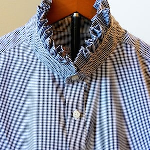 May include: A blue and white gingham button-down shirt featuring a high, ruffled stand collar. The shirt has a central placket with visible white buttons and a small partial label visible inside the collar area. The fabric pattern is a small, uniform check.