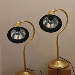 May include: Two brass desk lamps with black shades and curved necks. The lamps have a circular base and are positioned on wooden blocks. The lampshades have a silver interior and are connected to a power source via black cords.