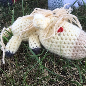 May include: A crocheted white horse with a red eye, lying on a bed of green grass.