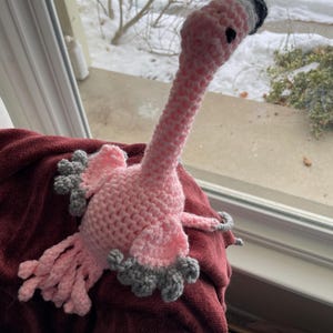 May include: A handmade pink crochet flamingo with gray accents on the wings and feet. The flamingo has a long neck and a black and white beak. The craft project is sitting on a burgundy fabric.
