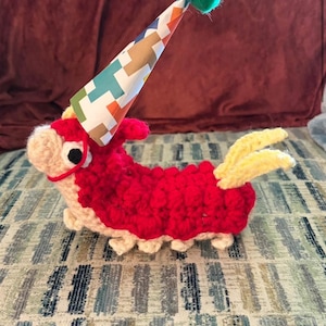 Pode incluir: Um peluche de croché que representa uma personagem Wurmple com um corpo vermelho texturado, cabeça e pés em bege claro e pontas de cauda amarelas. A criatura usa um chapéu de festa cónico com um padrão geométrico e um pompom verde.