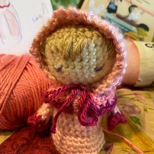 Peut inclure: Une poupée en crochet portant une robe à capuche rose avec un nœud rose et bordeaux. La poupée a les cheveux en laine brun clair et les yeux bleus.