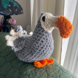 Könnte beinhalten: Handgehäkelter Dodo-Vogel in Grau-, Weiß- und Orangetönen. Der Vogel hat einen großen orangefarbenen Schnabel, einen weißen Kopf und einen grauen Körper. Die Füße sind orange. Der Dodo sitzt auf einer strukturierten grünen Oberfläche.