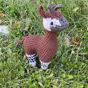 May include: A brown and white crocheted stuffed animal resembling a gerenuk, an antelope with a long neck, standing in a bed of green grass.