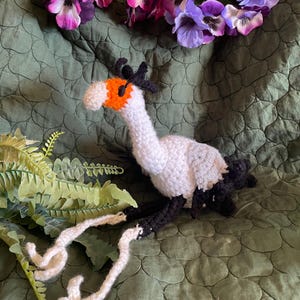 Pode incluir: Um brinquedo de pássaro de crochê feito à mão, com corpo branco, asas e pernas pretas e bico laranja. O pássaro está em uma superfície verde estampada, com um arranjo floral de flores roxas e rosas ao fundo.