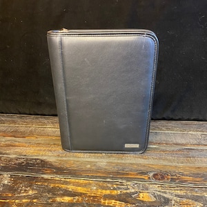 May include: A black leather portfolio with a zipper closure. The portfolio is rectangular and has a small silver-colored emblem on the lower right corner. It sits on a weathered wooden surface.