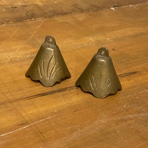 May include: Two small, antique brass bells with a floral design etched into the sides. Each bell has a loop at the top for hanging. The bells are set on a wooden surface.