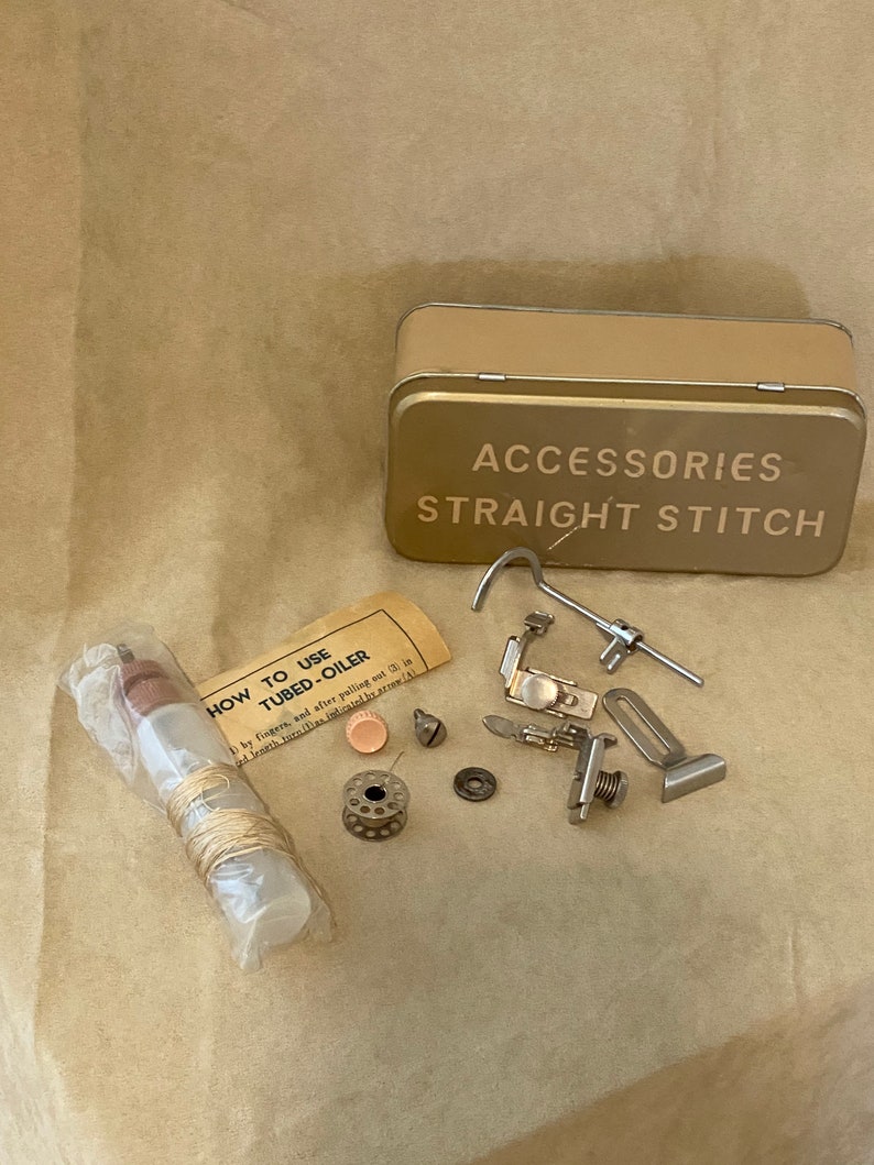 May include: A vintage sewing machine accessory kit in a tin box with the words "Accessories Straight Stitch" printed on the lid. The kit includes a variety of metal parts, a spool of thread, a small bottle of oil, and instructions on how to use the oil.
