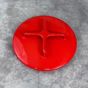 May include: A circular, bright red enameled lid featuring a raised, cross-shaped handle centered on the top surface. The lid has a smooth, glossy finish and a uniform, vibrant red color throughout its entire exterior surface.