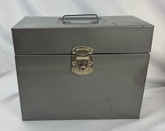 May include: A gray metal storage box with a handle and a latch closure. The box is rectangular and has a textured surface. The latch is silver-colored and has the words "Master Lock" on it.