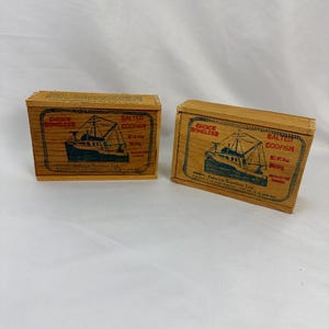 May include: Two wooden boxes of salted codfish. Each box has a green and gold label with the words "Choice Boneless Salted Codfish" and a drawing of a ship. The boxes are made of light brown wood.