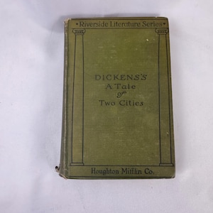 May include: A vintage copy of "A Tale of Two Cities" by Charles Dickens. The book has a green cover with black text and decorative columns. The title and author are clearly visible, along with the publisher, Houghton Mifflin Co. The book is part of the Riverside Literature Series.