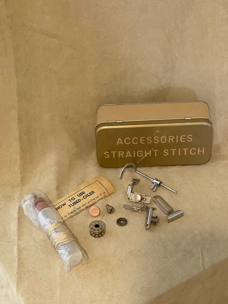 May include: A gold metal tin with the words "ACCESSORIES STRAIGHT STITCH" printed on the lid. The tin contains various sewing machine accessories, including a bobbin, a needle, a small metal tube, and a small piece of thread. There is also a small plastic bottle with a label that reads "HOW TO USE TUBED-OILER".