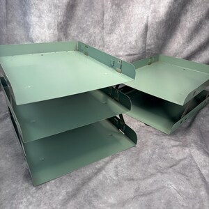 May include: A set of sage green metal document trays, including one three-tier stackable unit and one single-tier unit. Each tray features a flat base with raised sides and metal connectors at the corners for stacking, finished in a smooth matte paint.