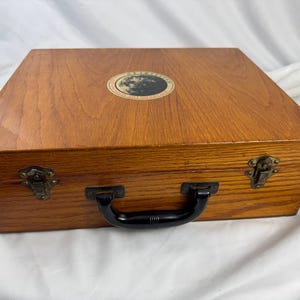 May include: A wooden box with a handle and metal clasps. The box is rectangular with a light brown finish and a circular emblem on the top. The handle is black, and the clasps are a dark metal color.