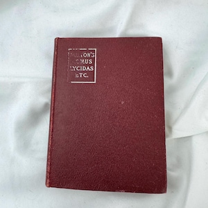 May include: A small, burgundy-colored book with the title "MILTON'S COMUS LYCIDAS ETC." in a white rectangular box on the upper left corner. The book has a textured cover and is set against a white background.