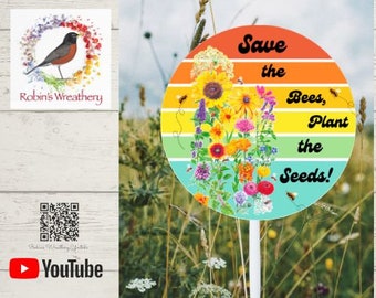 May include: A colourful round garden sign with a rainbow background and a message that reads "Save the Bees, Plant the Seeds!" The sign features a variety of flowers and bees.