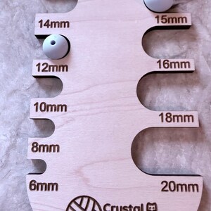 Pu&ograve; includere: Un misuratore di perline in legno con la scritta "BEAD SIZER" e misure da 6 mm a 20 mm. Due perline bianche sono posizionate nel misuratore. &Egrave; visibile anche la scritta "Crystal Makes the Things".