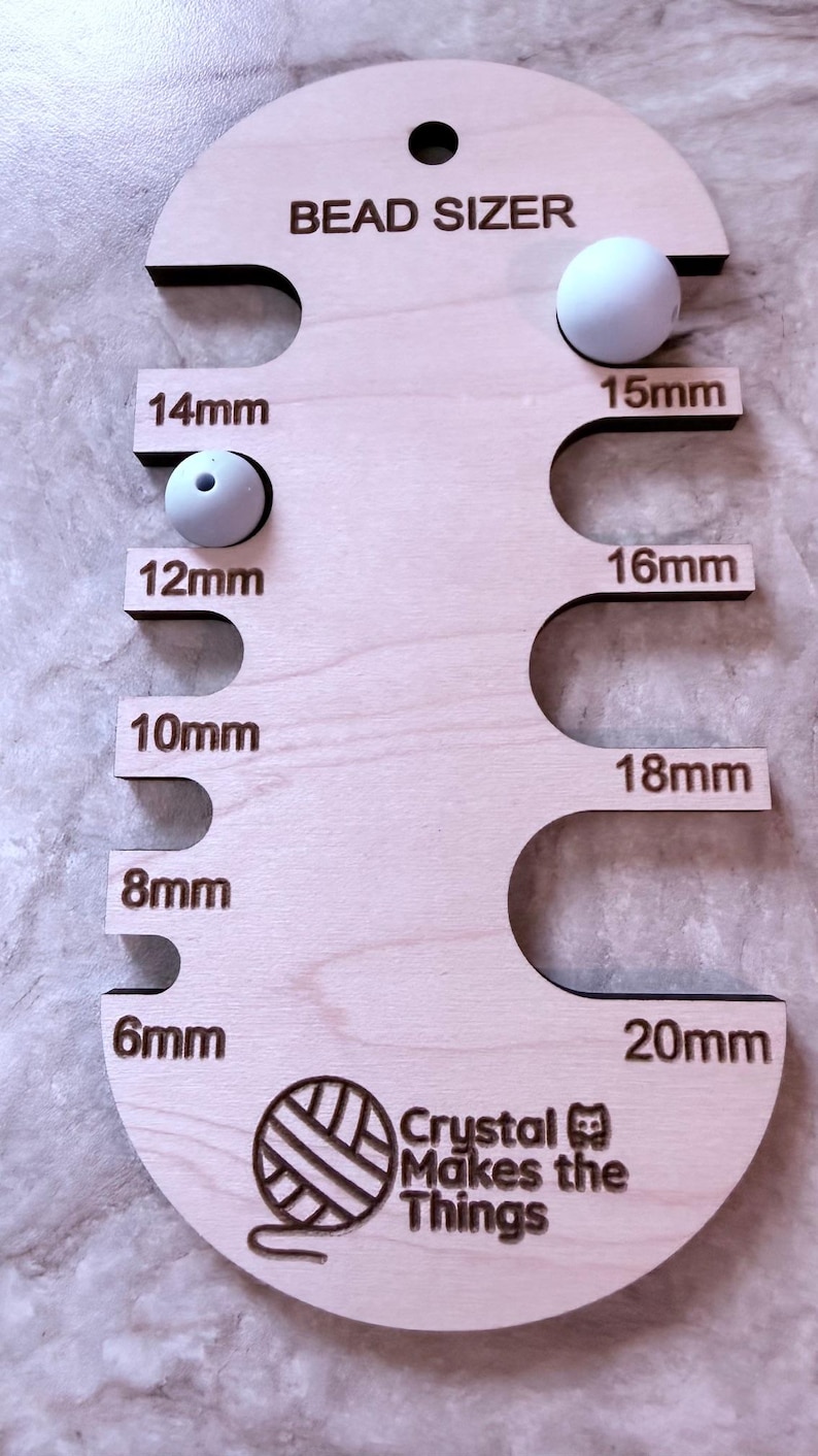 Pu&ograve; includere: Un misuratore di perline in legno con la scritta "BEAD SIZER" e misure da 6 mm a 20 mm. Due perline bianche sono posizionate nel misuratore. &Egrave; visibile anche la scritta "Crystal Makes the Things".