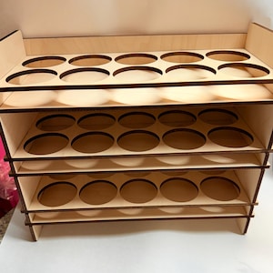 Puede incluir: Una estantería de madera con tres niveles y una balda superior. Cada nivel tiene recortes circulares para sujetar objetos. La madera tiene un color claro natural con bordes más oscuros, y la estantería está diseñada para estar de pie.