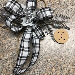 May include: A black and white plaid ribbon bow with silver glitter and beads, tied to a brown twine string. The bow is decorated with a silver fern sprig. A brown paper circle with six holes is in the background.