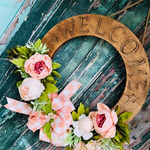 Könnte beinhalten: Ein Holz-Kranz mit dem Wort "WELCOME" darauf geschnitzt. Der Kranz ist mit rosa und weißen Blumen und einer pfirsichfarbenen Gingham-Schleife verziert.