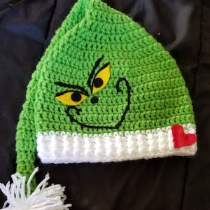 May include: Green crocheted hat with a Grinch face and a red heart on the side. The hat has a white pom-pom on the end of the long tail.