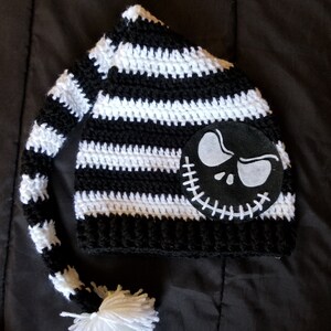 May include: Black and white striped crochet hat with a long tail and a white pom-pom. The hat features a black felt applique of Jack Skellington's face from The Nightmare Before Christmas.
