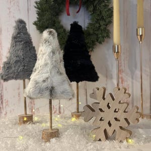 May include: A festive holiday display featuring three faux fur Christmas trees in gray, white, and black, each mounted on a wooden base. A wooden snowflake and candles complete the winter scene, with a green wreath in the background.
