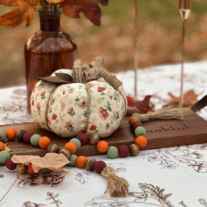 May include: A fabric pumpkin with a floral pattern, twine stem, and leaf accents sits on a wooden board. A wooden bead garland in fall colors and a brown glass bottle with fall leaves complete the autumn-themed display.